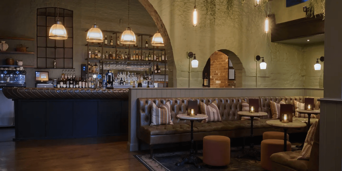 Dimly lit bar interior with leather banquette seating, pendant lights and shelved spirits bottles.