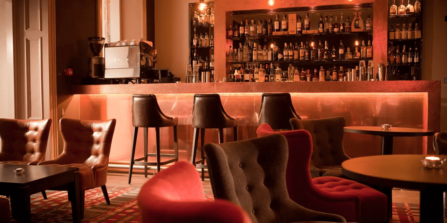 Dimly lit hotel bar with leather armchairs, bar stools and shelves of spirits bottles.