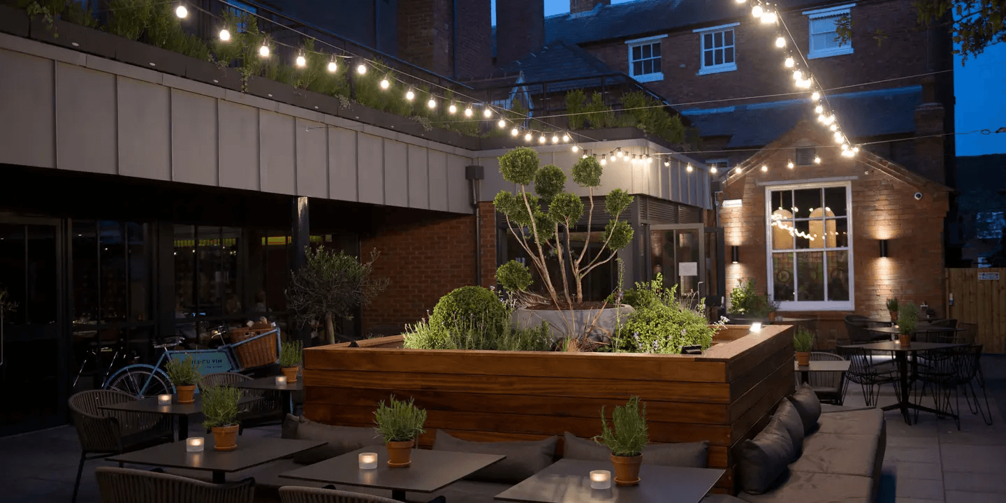 Courtyard terrace at dusk with festoon lights, a central planted bed and bistro tables.