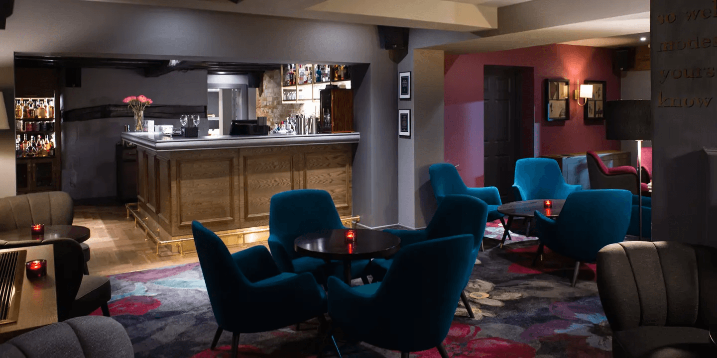 Dimly lit hotel bar with teal velvet armchairs, round tables and candlelight