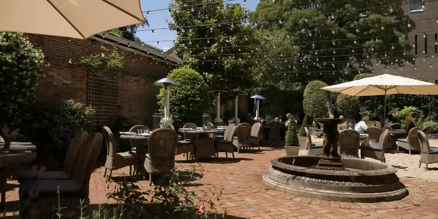 Sunny walled courtyard garden with rattan seating, parasols, festoon lights and a stone fountain.