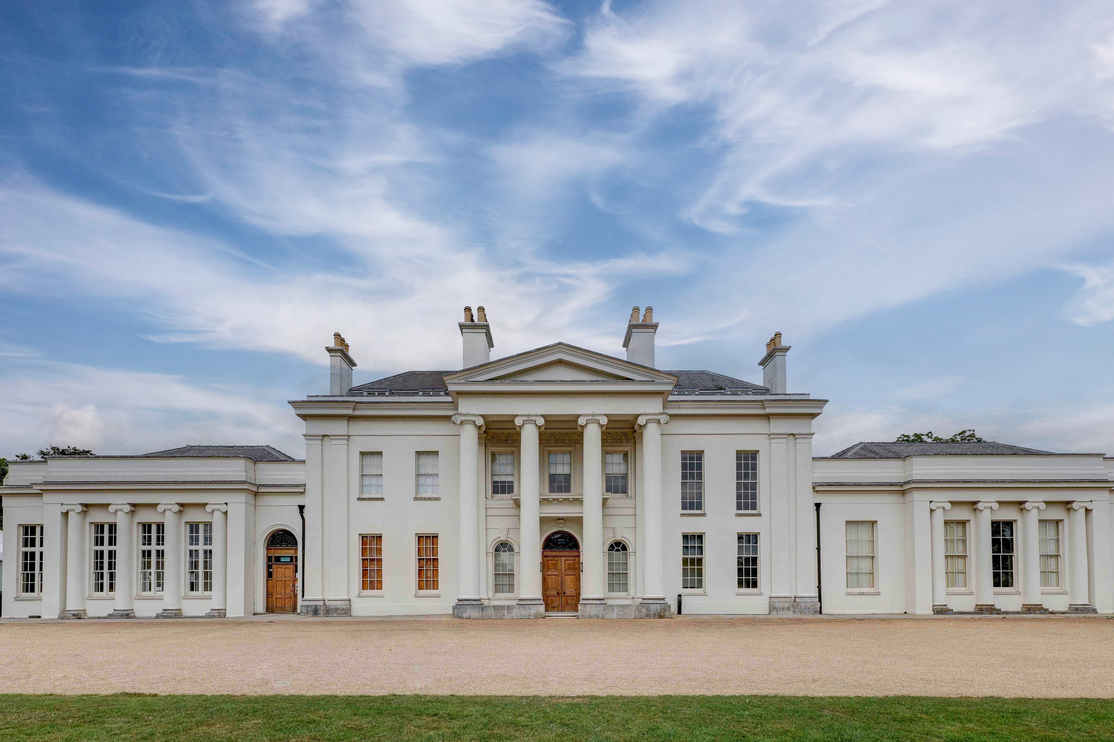 The white neoclassical exterior of Hylands House with columned portico and gravel forecourt.