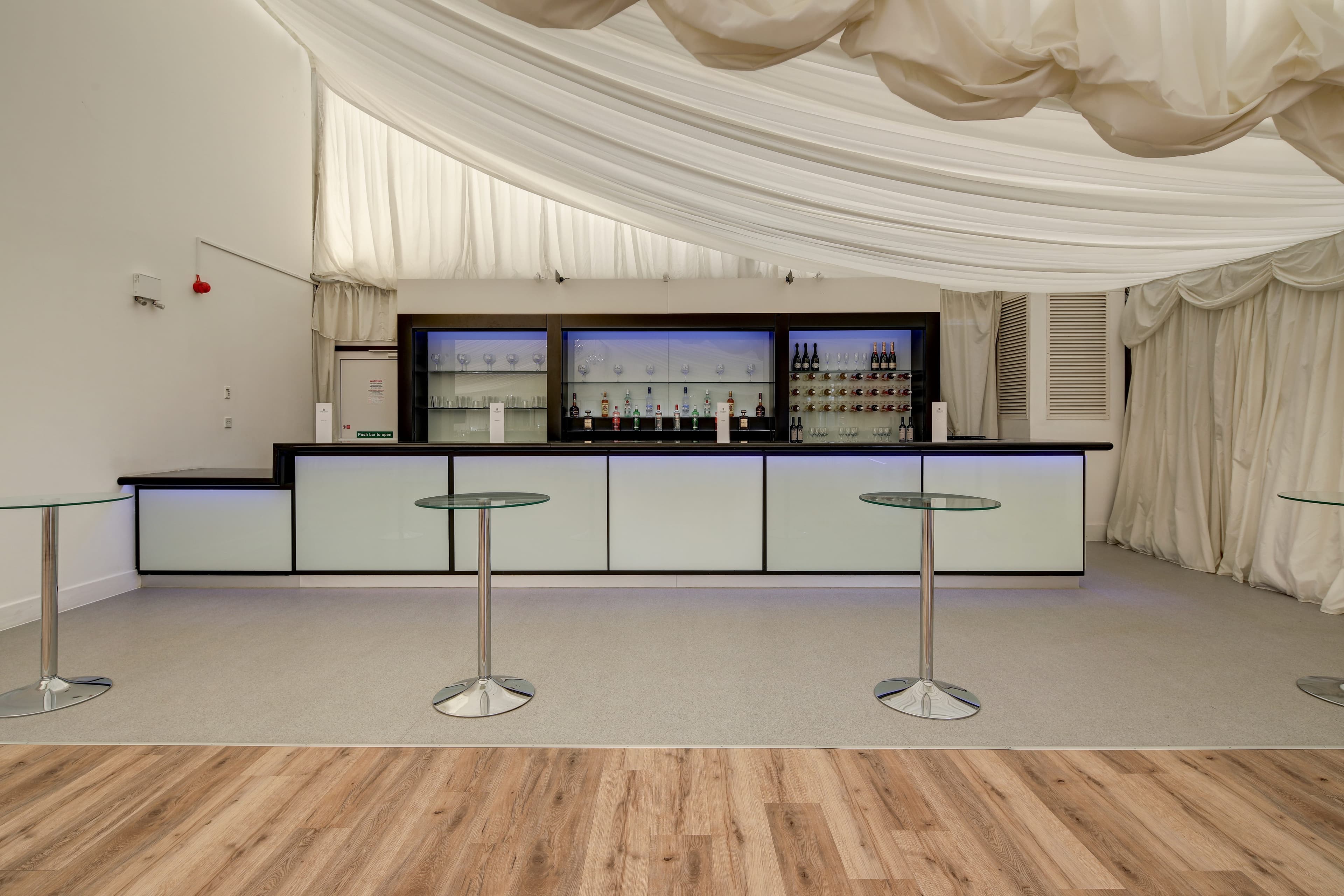 Illuminated bar with glass-topped poseur tables beneath white draped ceiling at Hylands Estate.