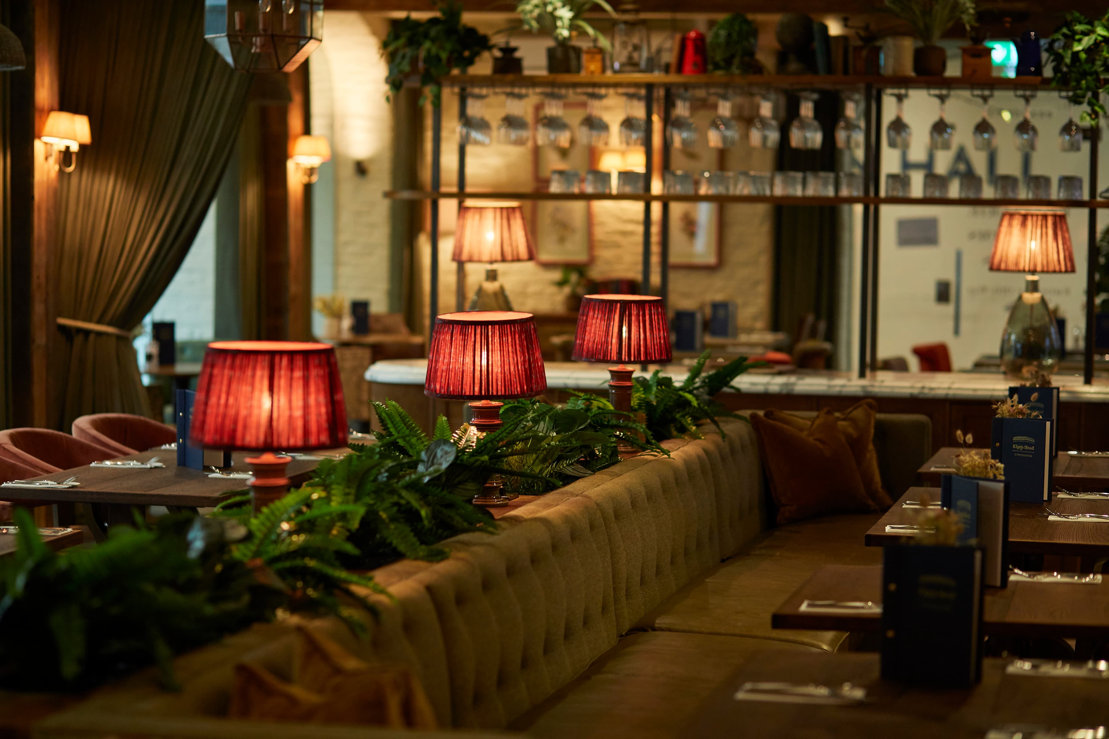 Dimly lit restaurant interior with red table lamps, banquette seating and fern garlands.