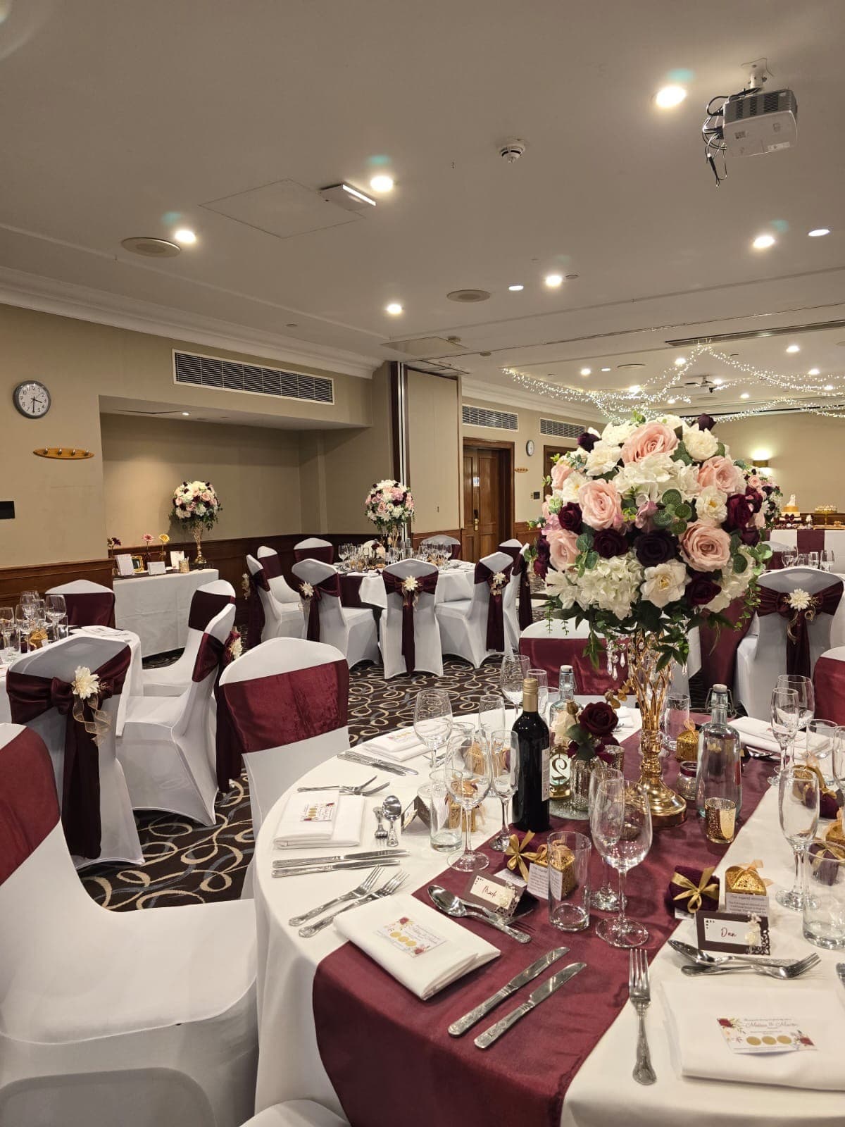 Banqueting room laid for a wedding with burgundy sashes and tall floral centrepieces.