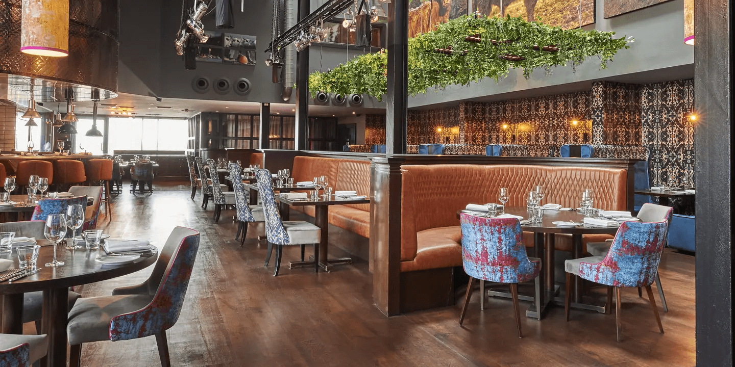 Restaurant interior with tan leather booths, patterned chairs and hanging greenery above.