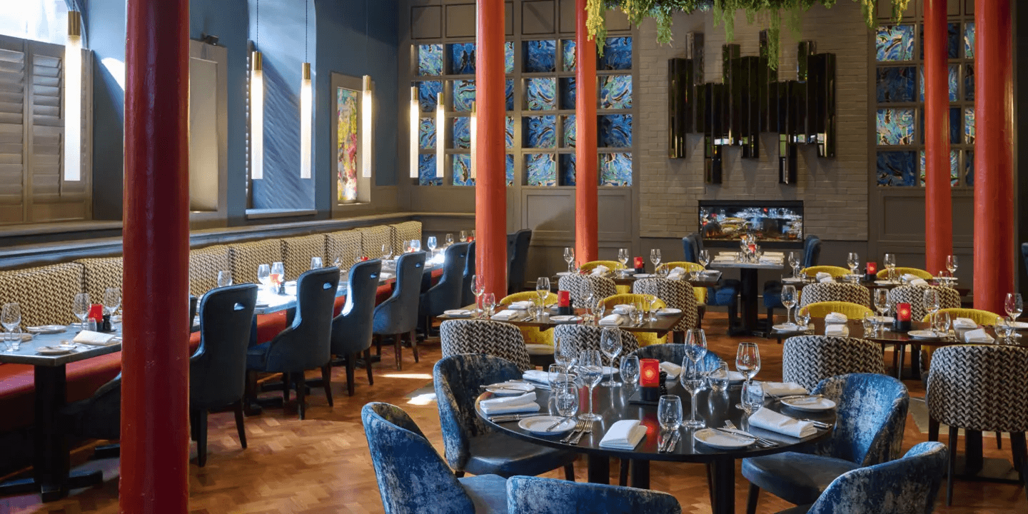 Restaurant dining room with round tables, velvet chairs and colourful stained glass panels.