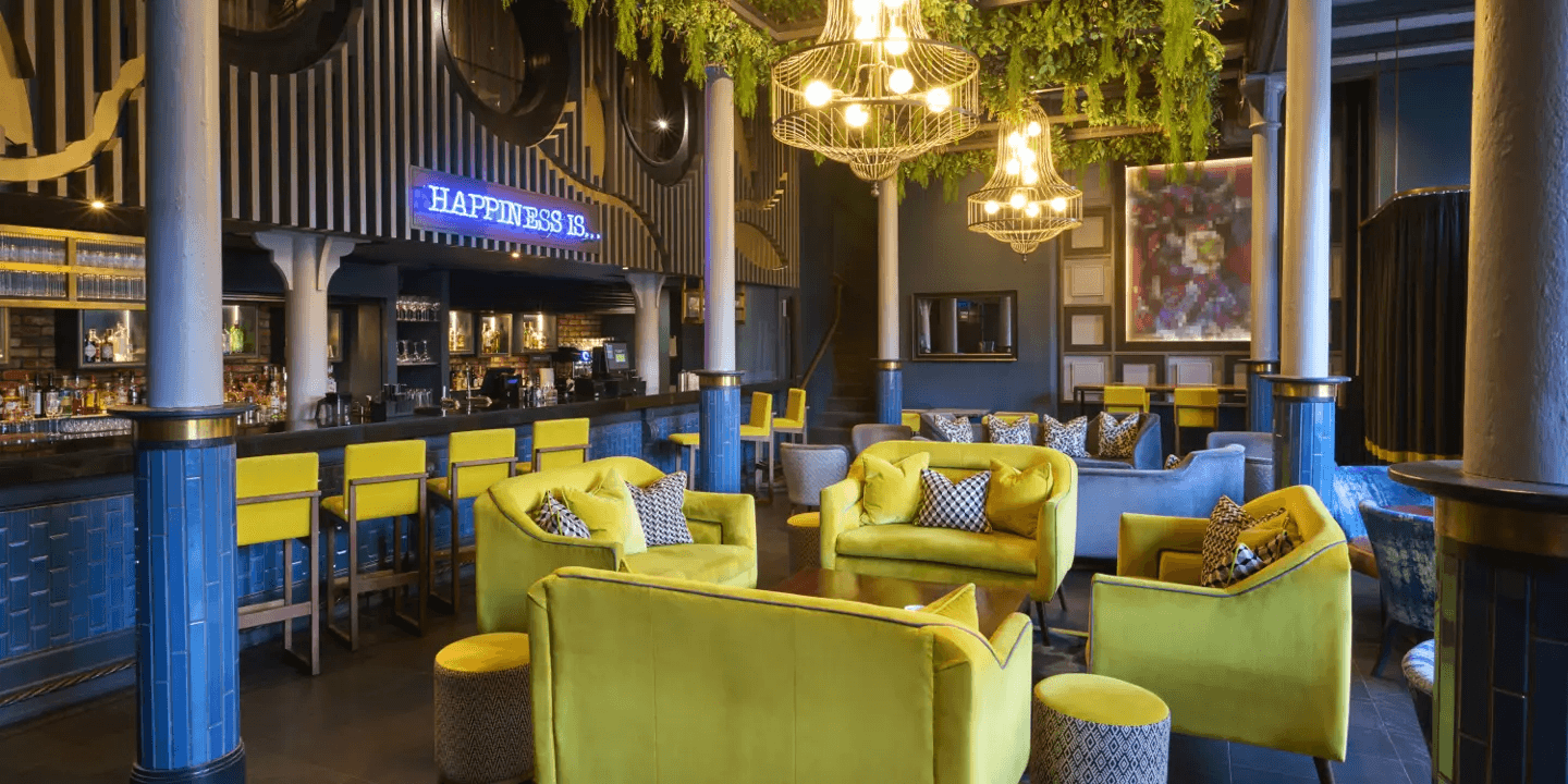 Stylish hotel bar with yellow velvet armchairs, blue tiled counter and a neon sign reading Happiness Is.