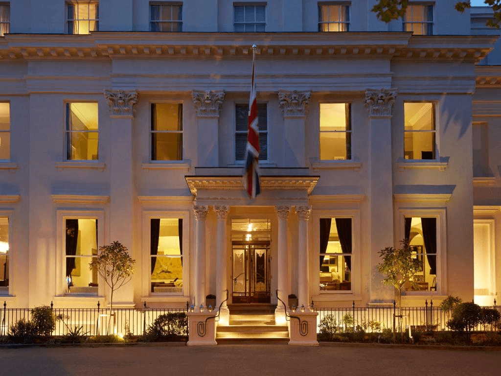The white stucco exterior of Malmaison Cheltenham hotel lit up at dusk with a Union Jack flag.