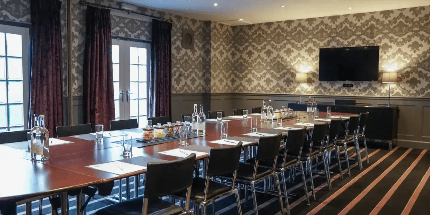 Boardroom-style meeting room with patterned wallpaper, wall-mounted screen and laid tables.