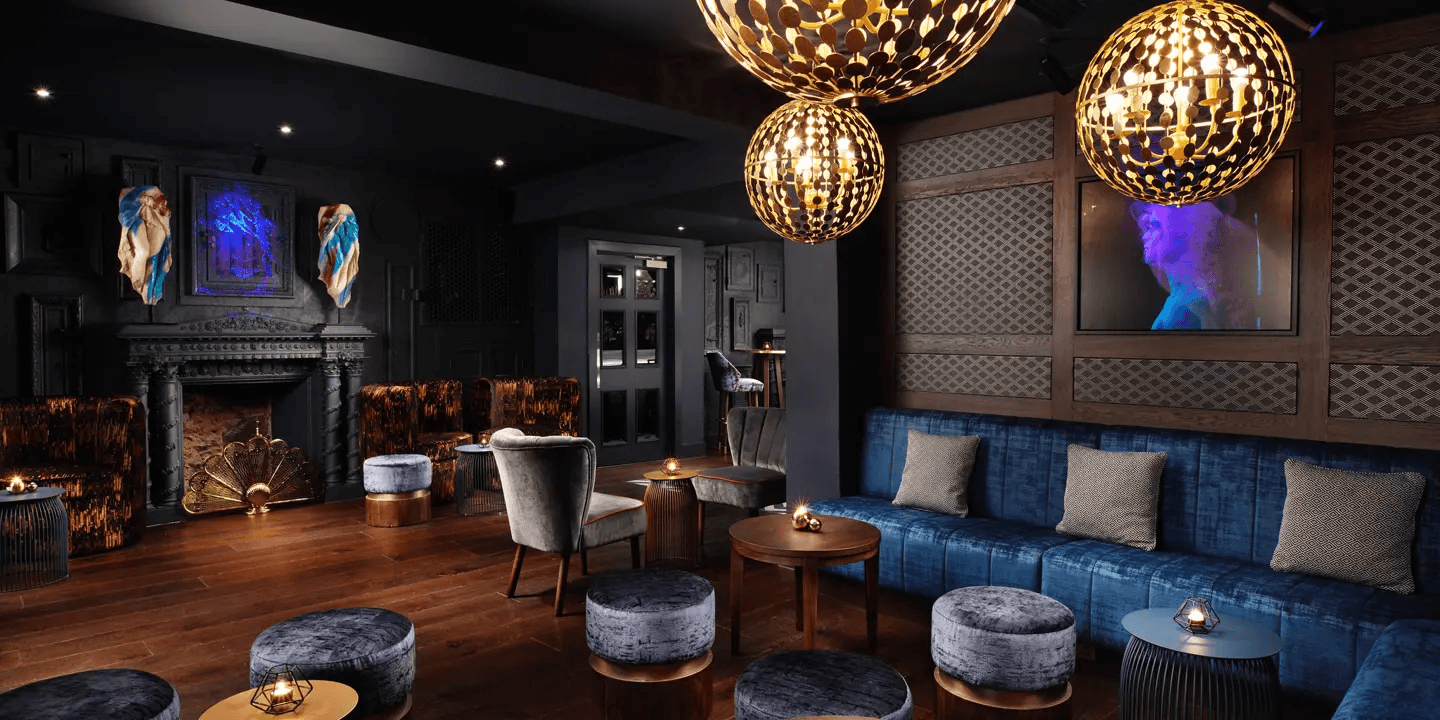 Moody cocktail bar with blue velvet sofas, globe pendant lights and an ornate fireplace.