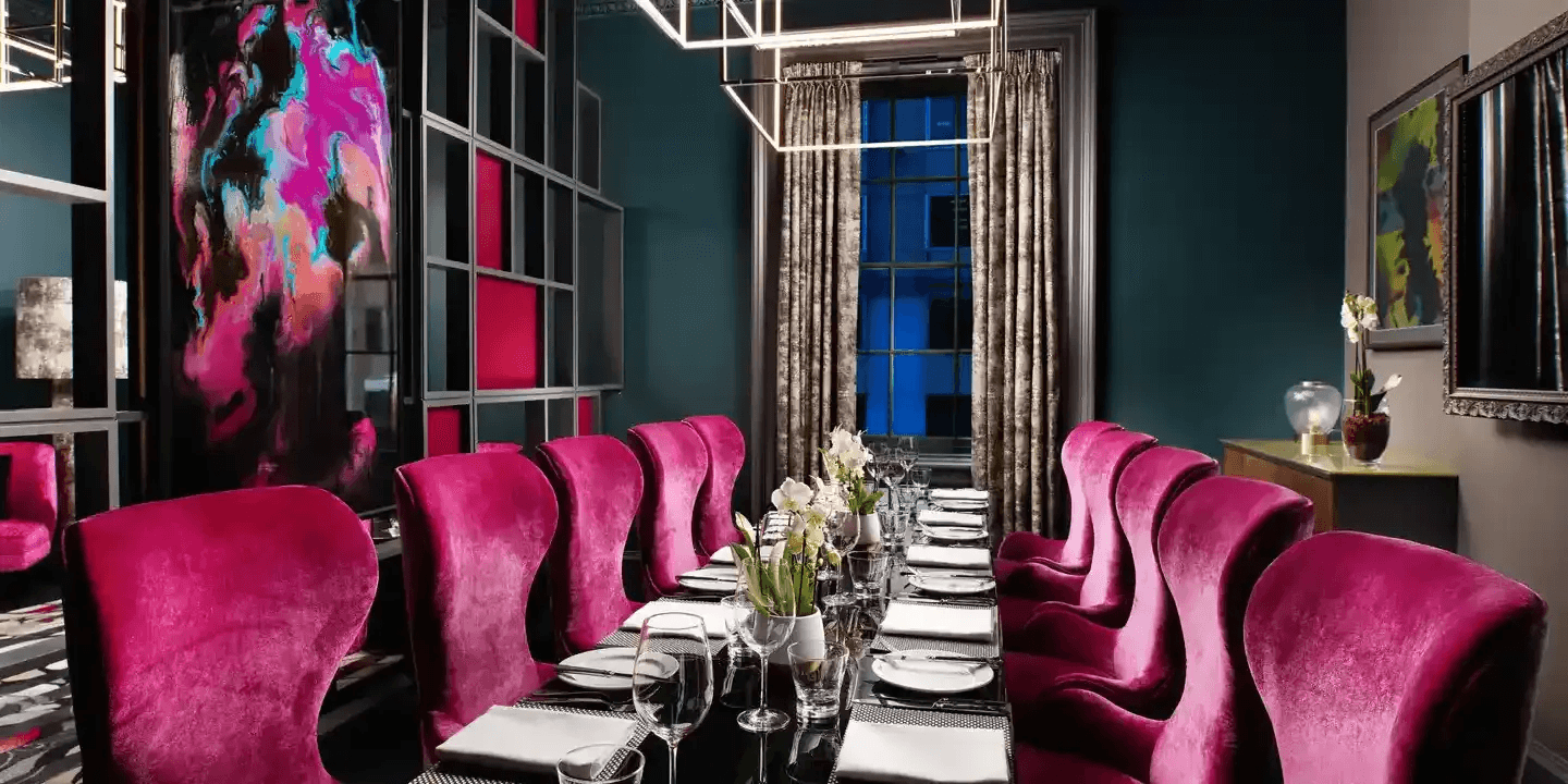Private dining room with magenta velvet chairs, a long set table and bold artwork.