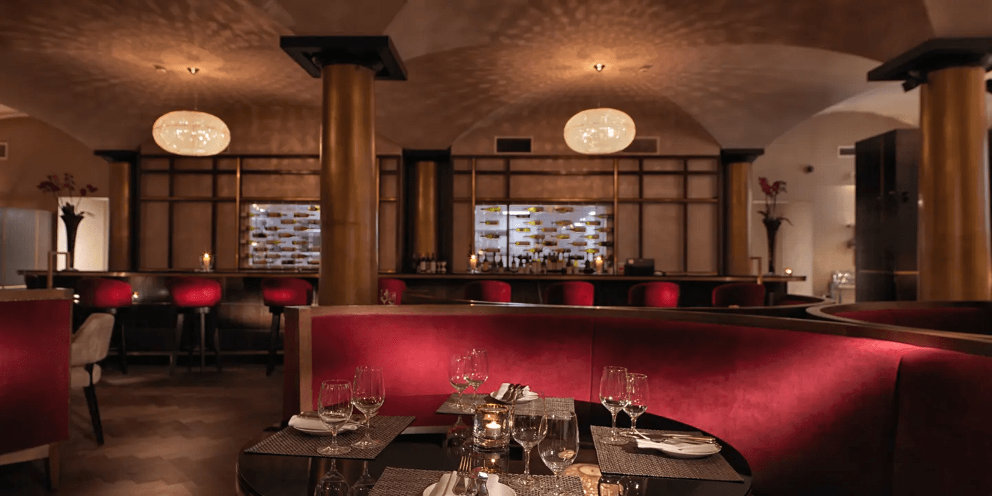 Dimly lit restaurant interior with red booth seating, bar and illuminated wine display behind.
