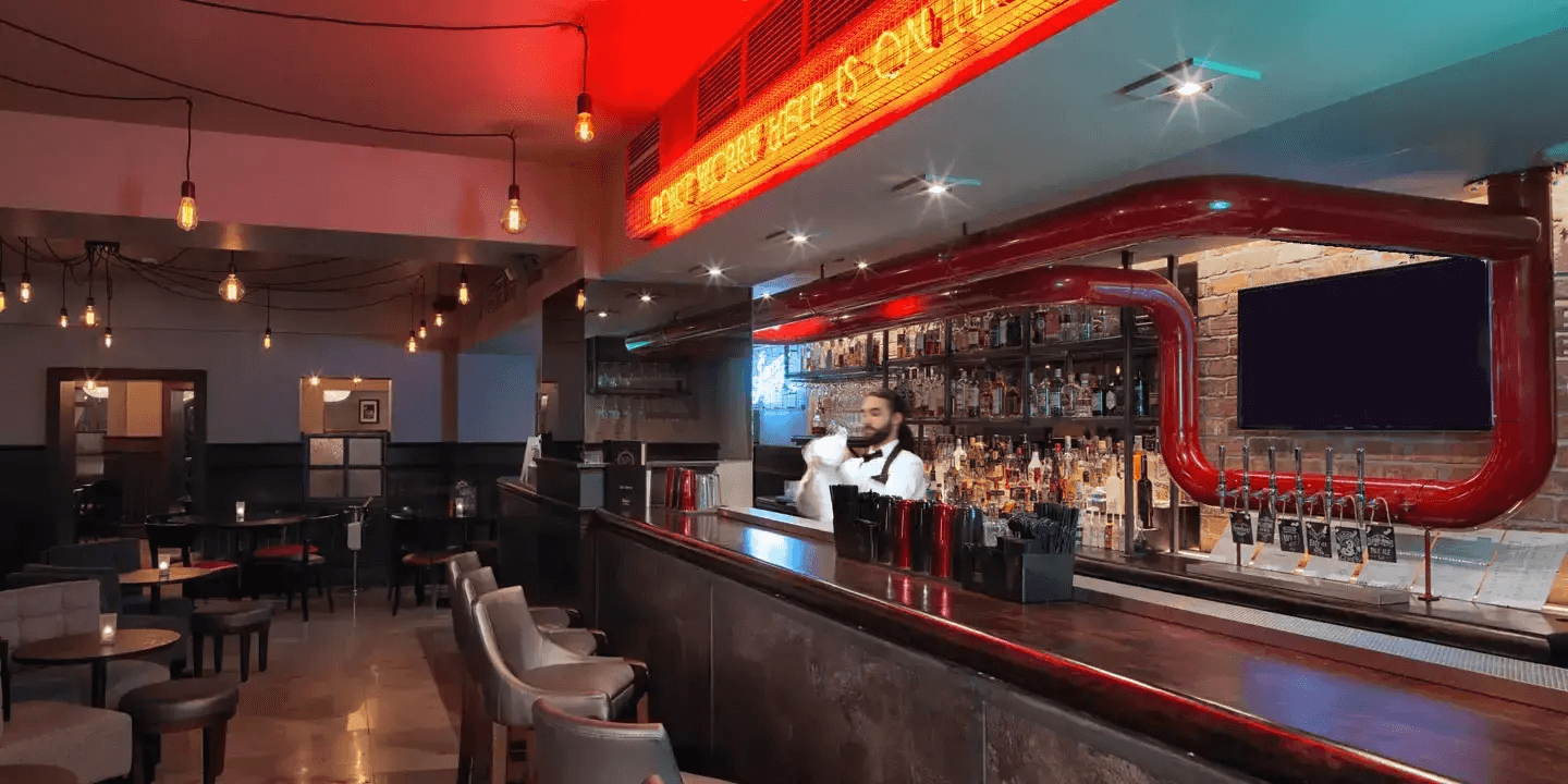 The dimly lit bar at Malmaison Leeds with a red neon sign overhead and a bartender working behind the counter.
