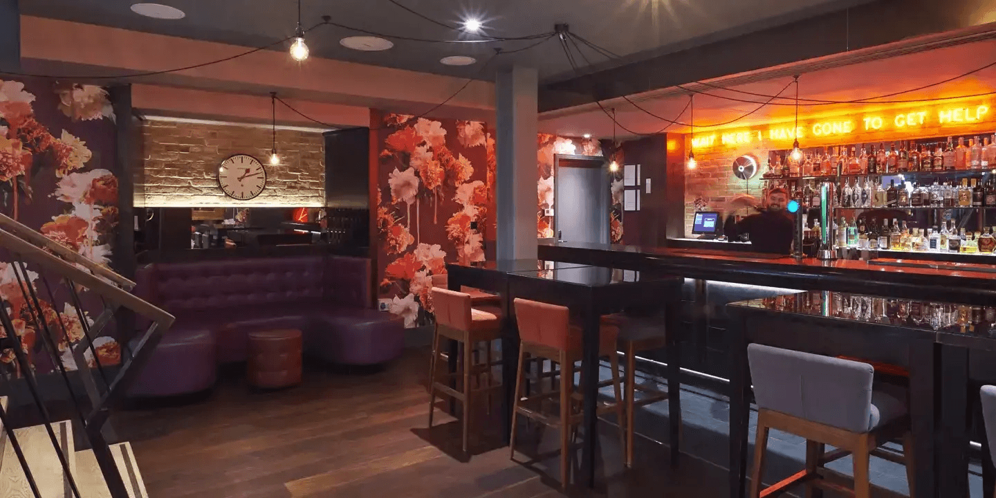 Dimly lit cocktail bar with floral wallpaper, purple booth seating and neon sign above shelves of spirits.