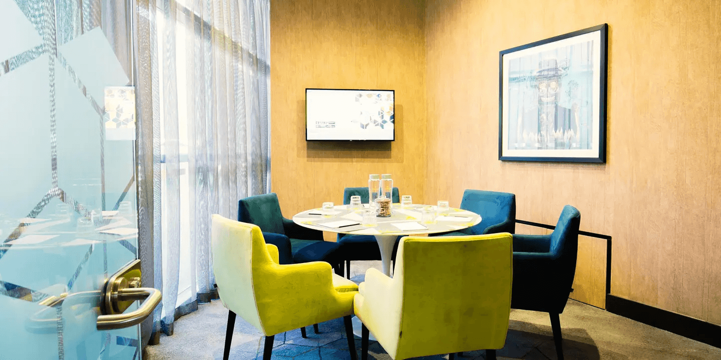 Small private meeting room with round table, yellow and teal chairs and wall-mounted screen.