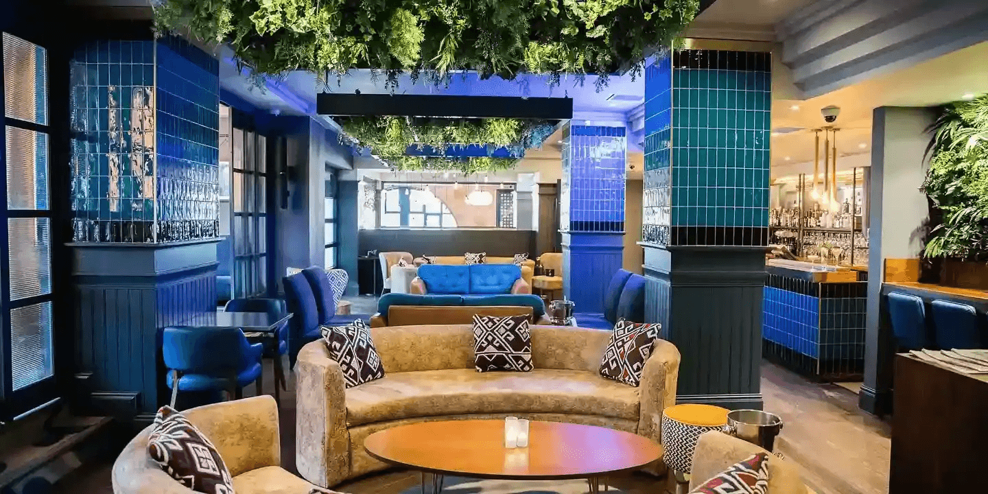 Cocktail bar lounge with curved sofas, teal tiled pillars and overhead foliage installation.