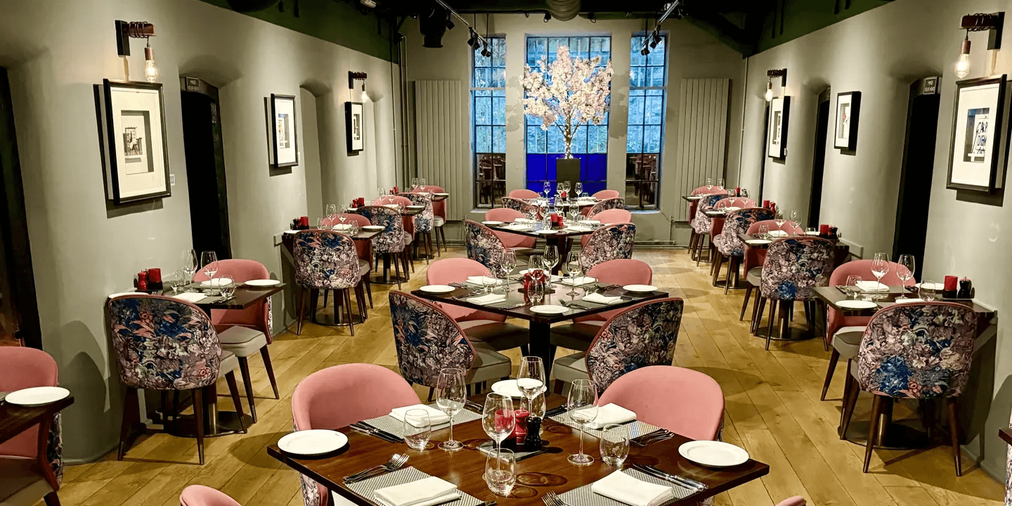 Restaurant dining room with pink and floral chairs, laid tables and a tall arched window.