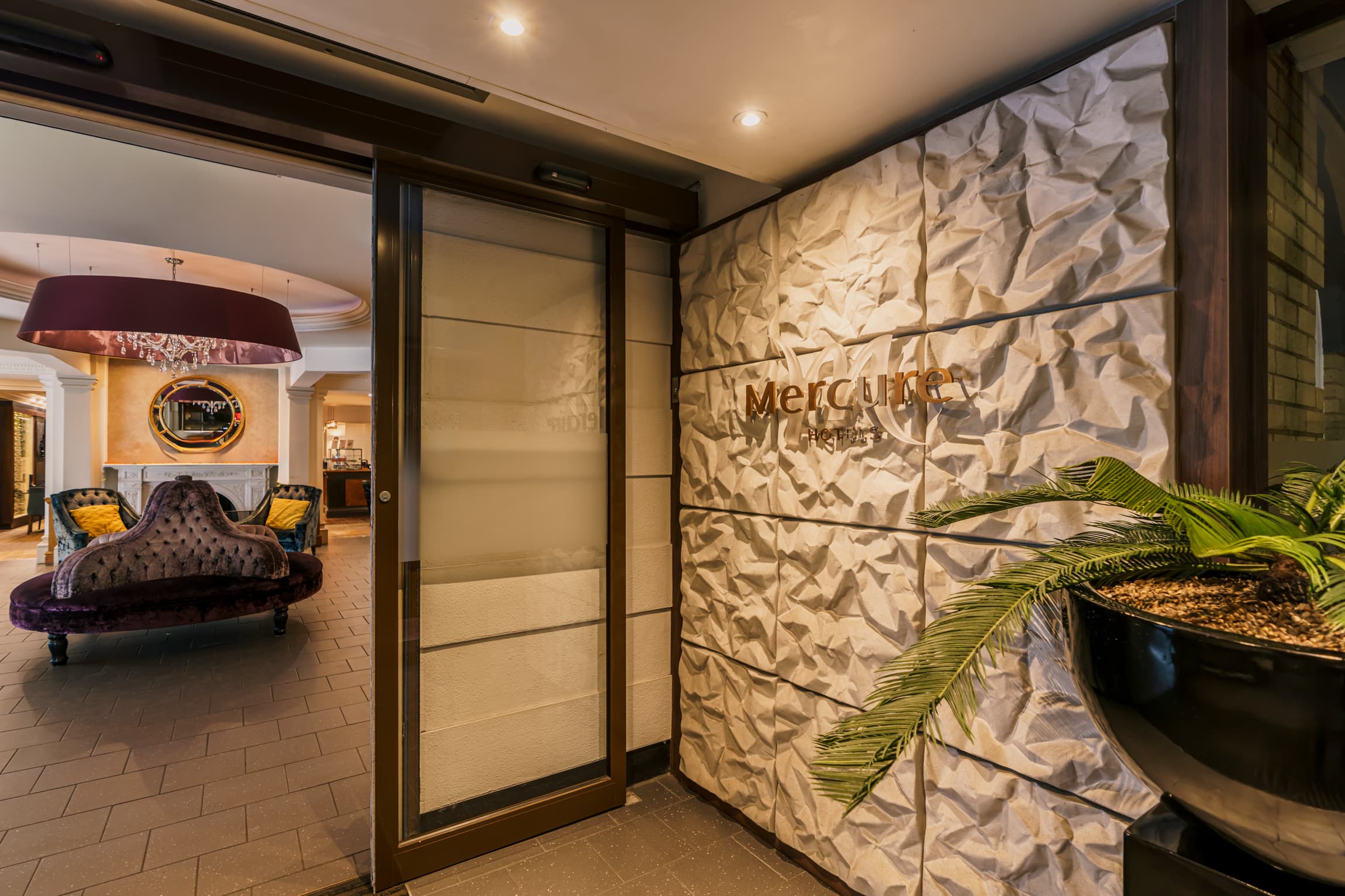 Hotel entrance with textured wall bearing the Mercure Hotels sign and a palm plant.