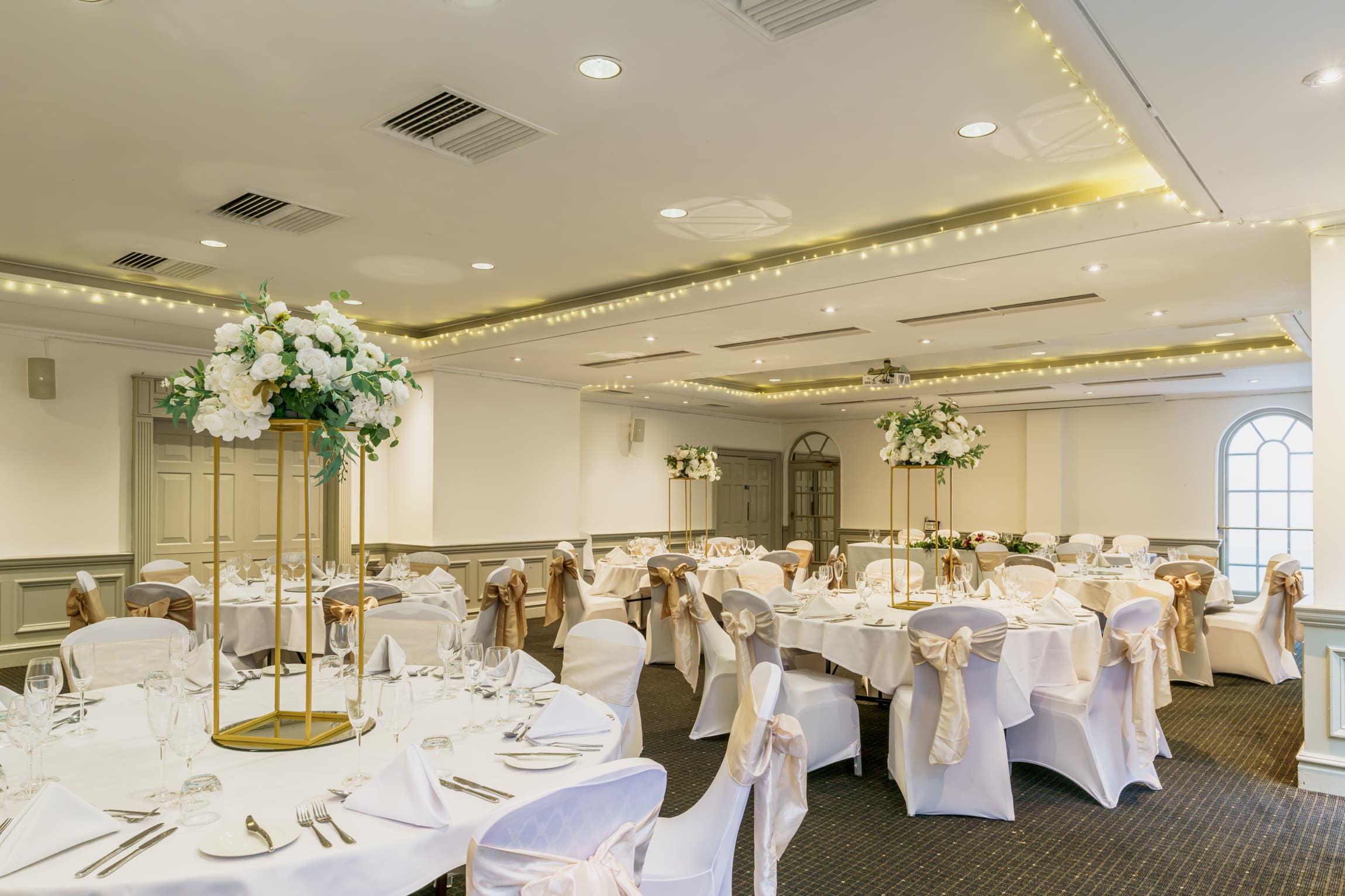 Banqueting room set for a wedding with round tables, white chair covers and tall floral centrepieces.