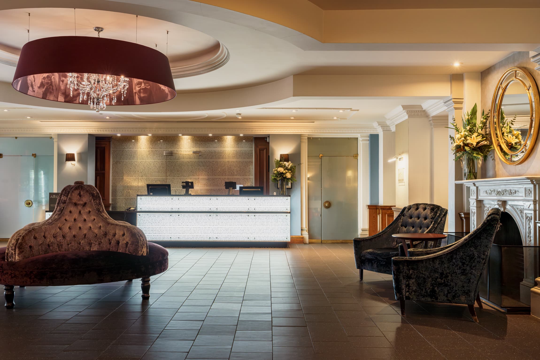 Hotel reception lobby with velvet seating, chandelier, ornate fireplace and front desk.