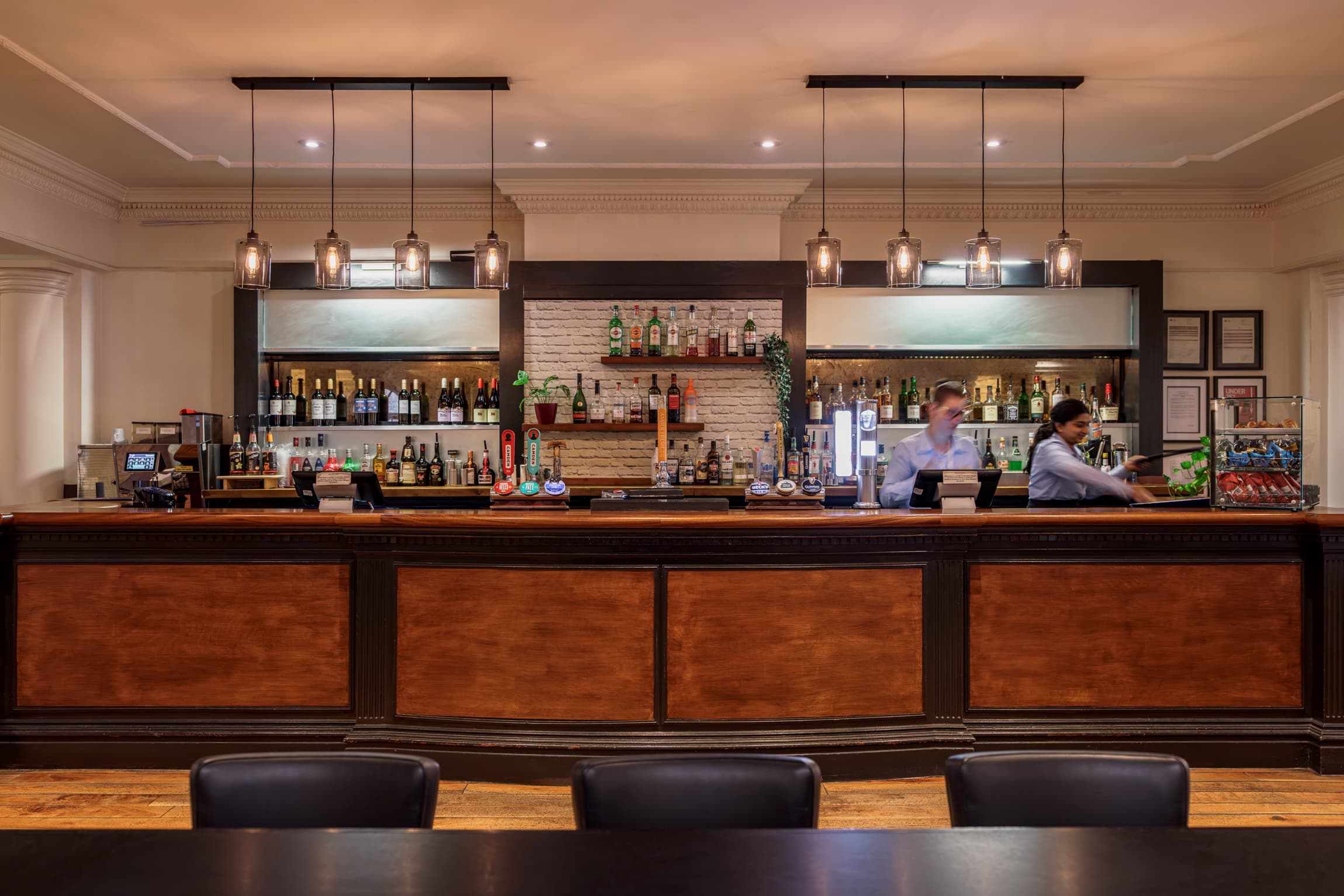 The hotel bar with timber panelling, draught taps and two staff members serving.