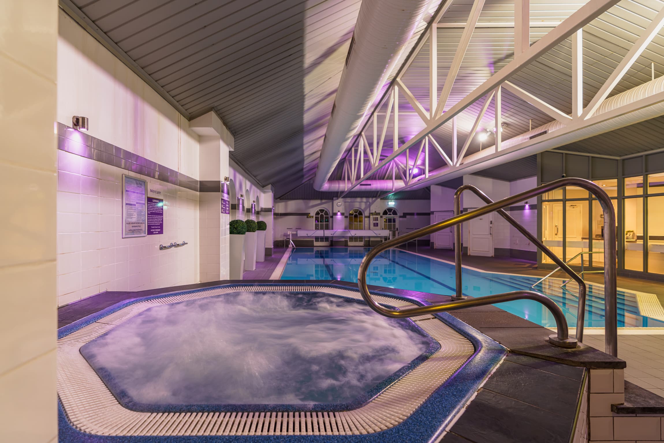 Indoor pool and bubbling hot tub under a vaulted ceiling with purple mood lighting.