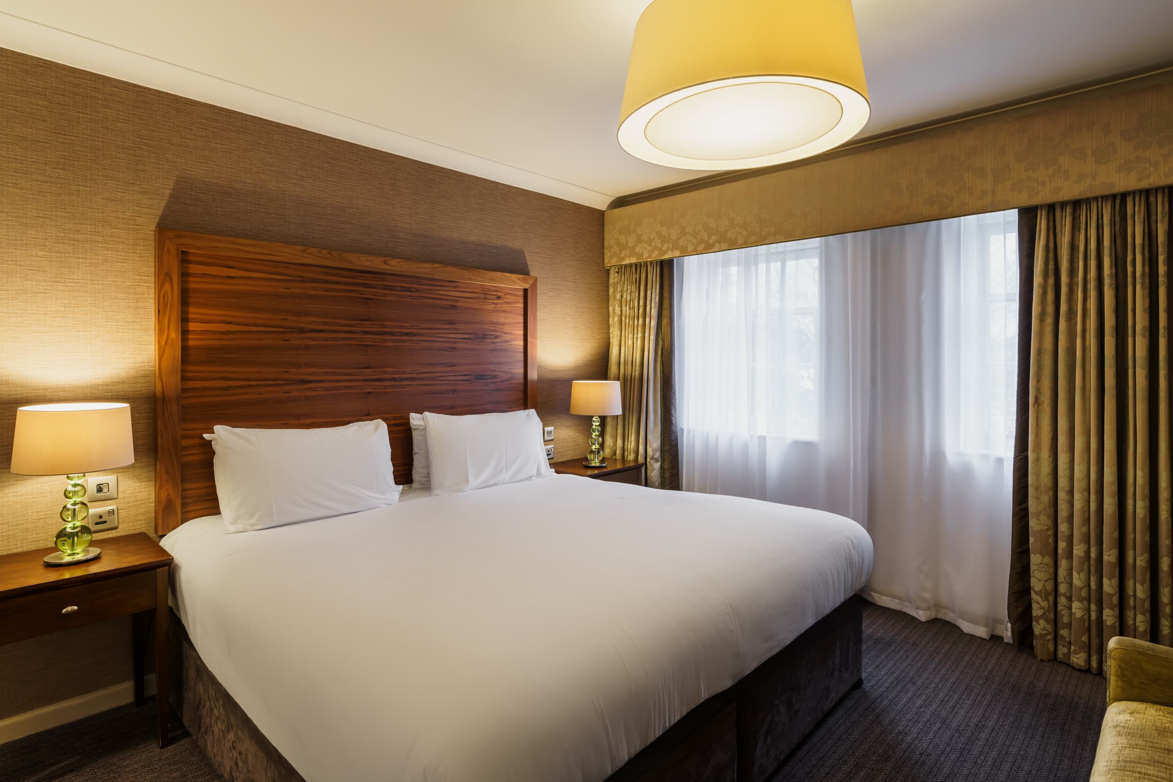 Double hotel room with a large wooden headboard, white bedding and gold curtains.