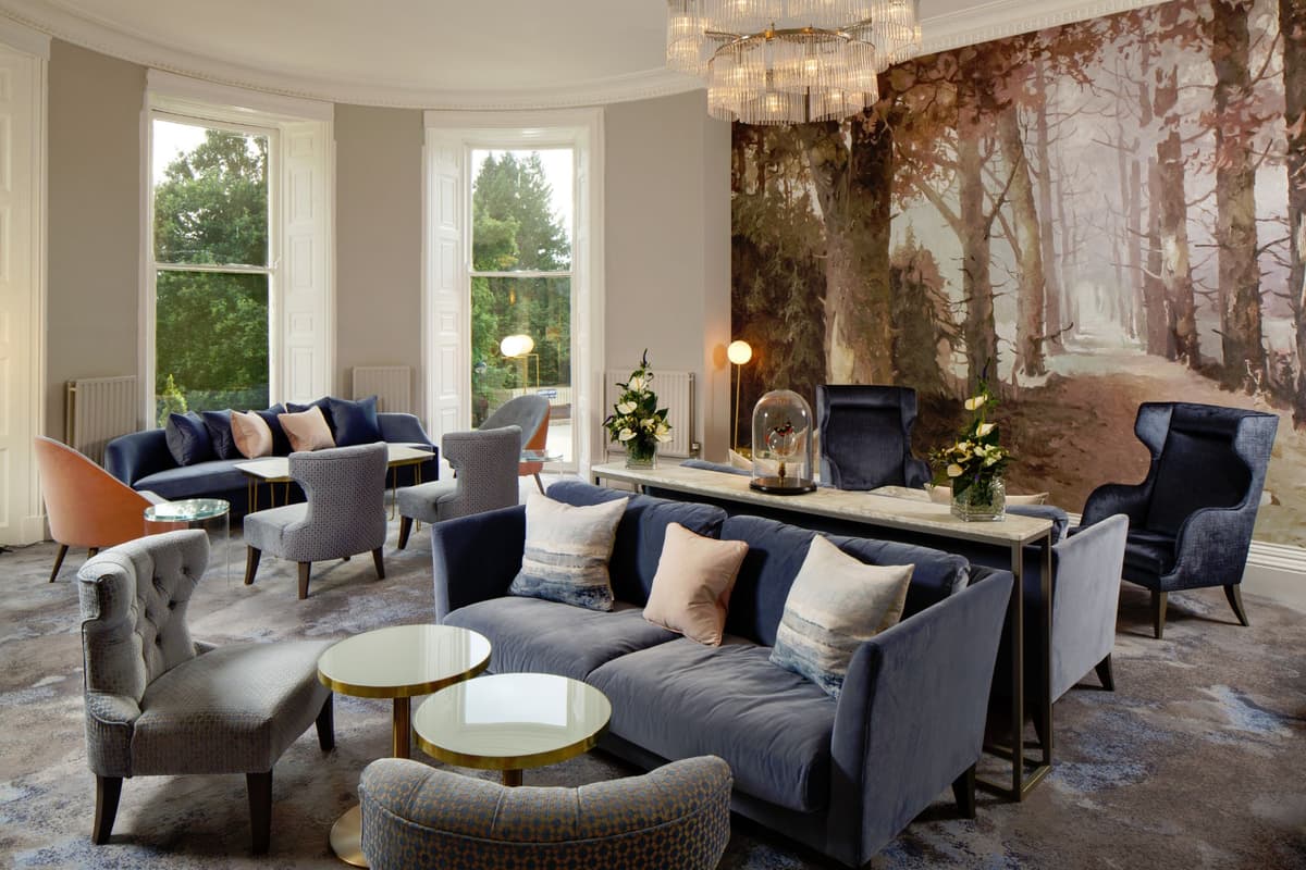 Elegant hotel lounge with blue velvet sofas, chandelier and woodland mural wallpaper.