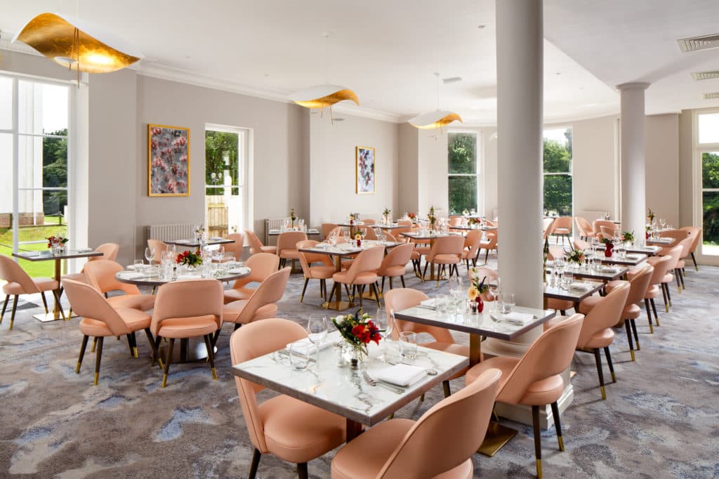 Bright hotel restaurant with pink chairs, marble tables and gold leaf pendant lights.