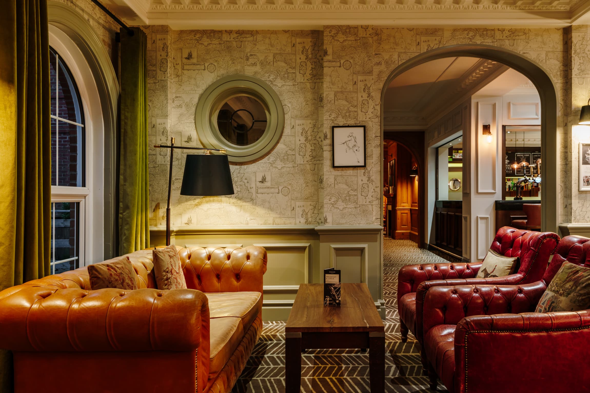 Leather Chesterfield sofas in a warm hotel lounge with map-print wallpaper and arched doorway.