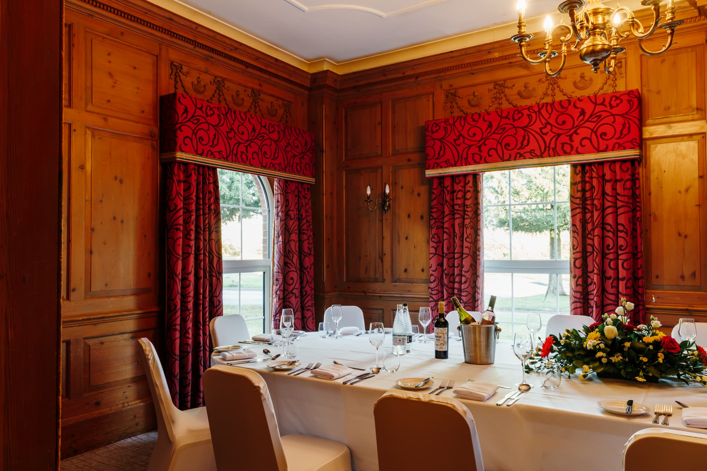 Wood-panelled private dining room with red curtains, laid table and floral centrepiece.