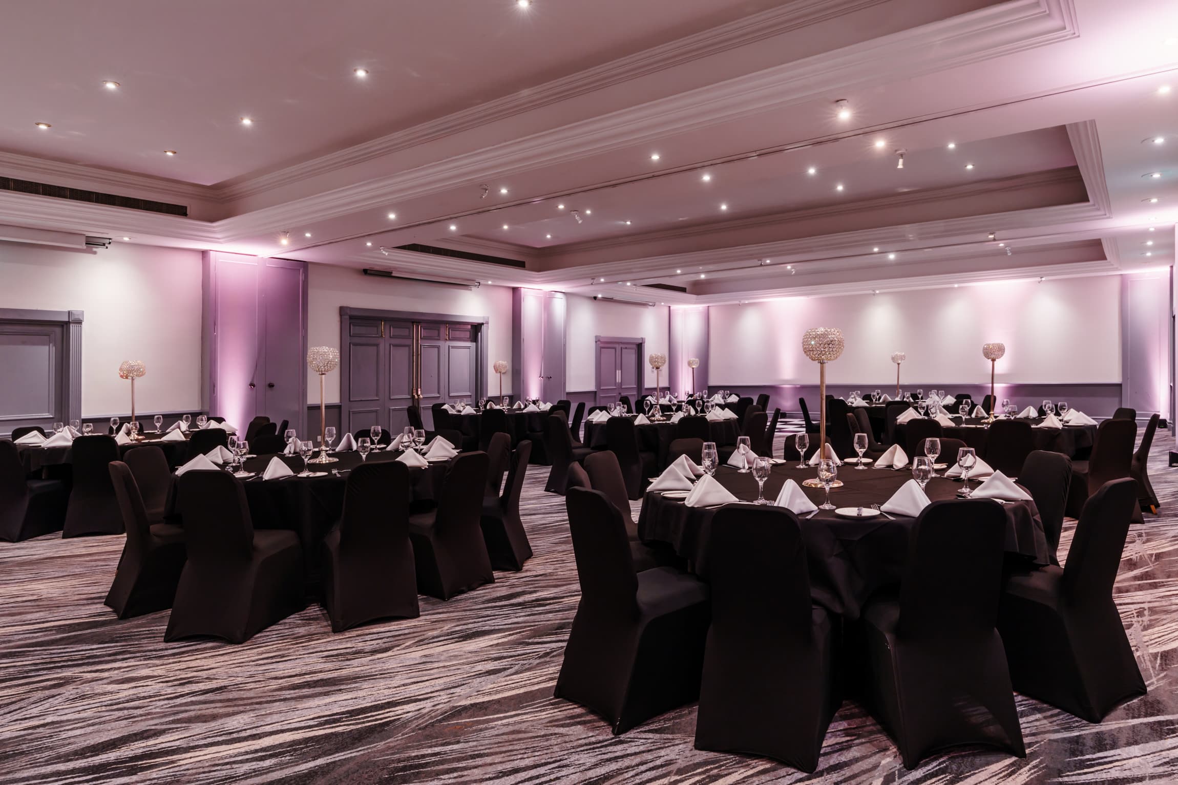 Ballroom laid for a banquet with round black-draped tables and pink uplighting.
