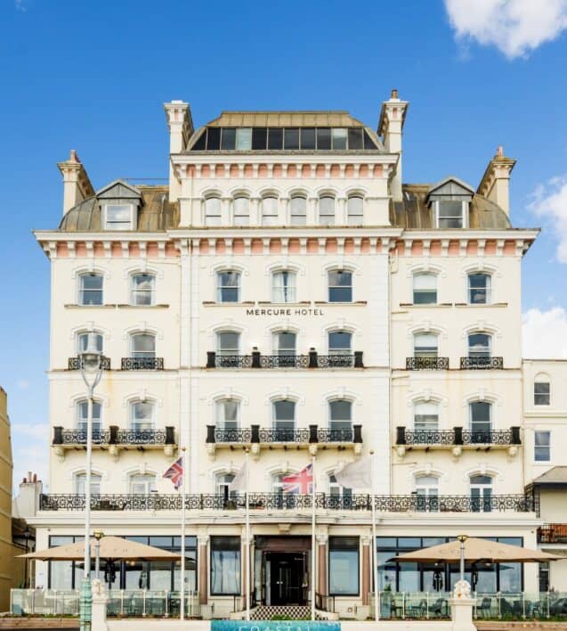 The white Victorian seafront facade of Mercure Hotel Brighton with ornate balconies and Union flags.