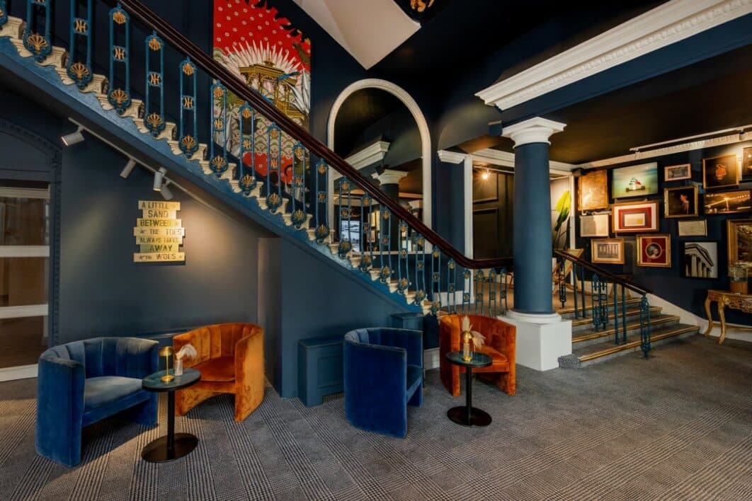 Hotel lobby with ornate blue staircase, velvet tub chairs and a gallery wall.