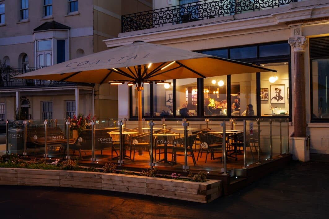 The Coastal Restaurant terrace at night with lit parasols and glass screens.