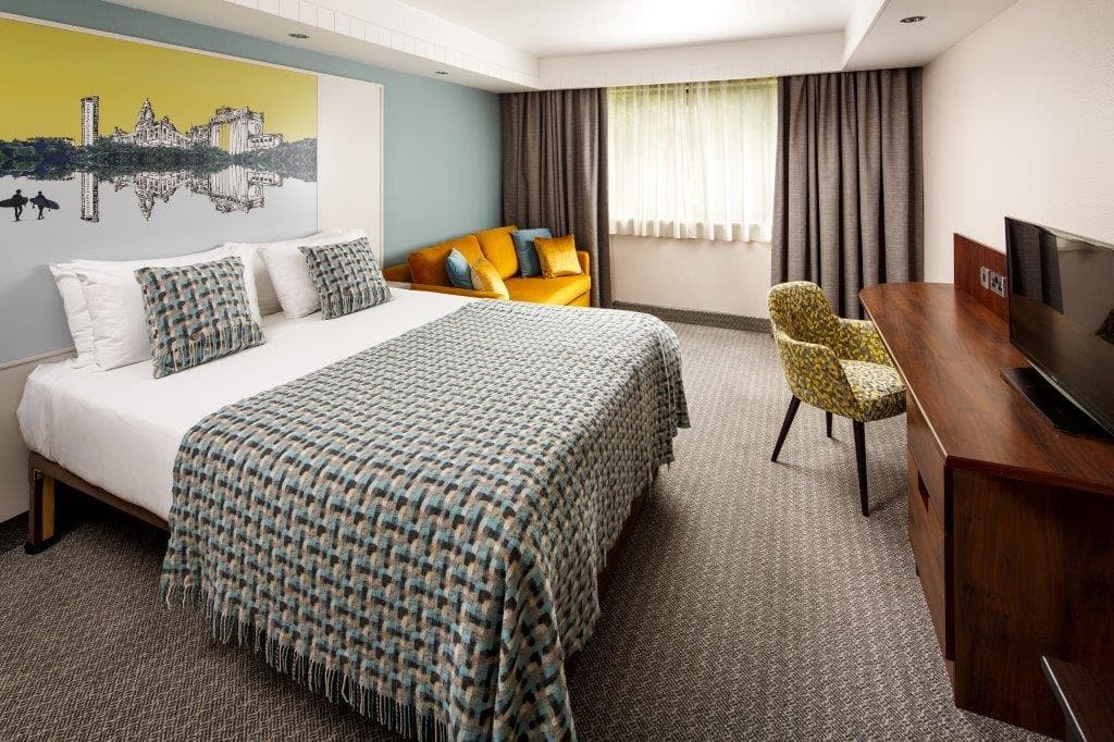 Hotel bedroom with double bed, mustard sofa, patterned throw and wood desk.