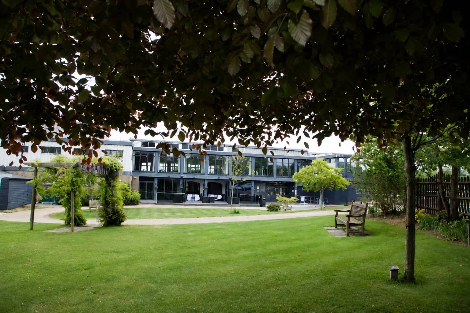 Manicured garden lawn with trees, a wooden bench and glazed building beyond.