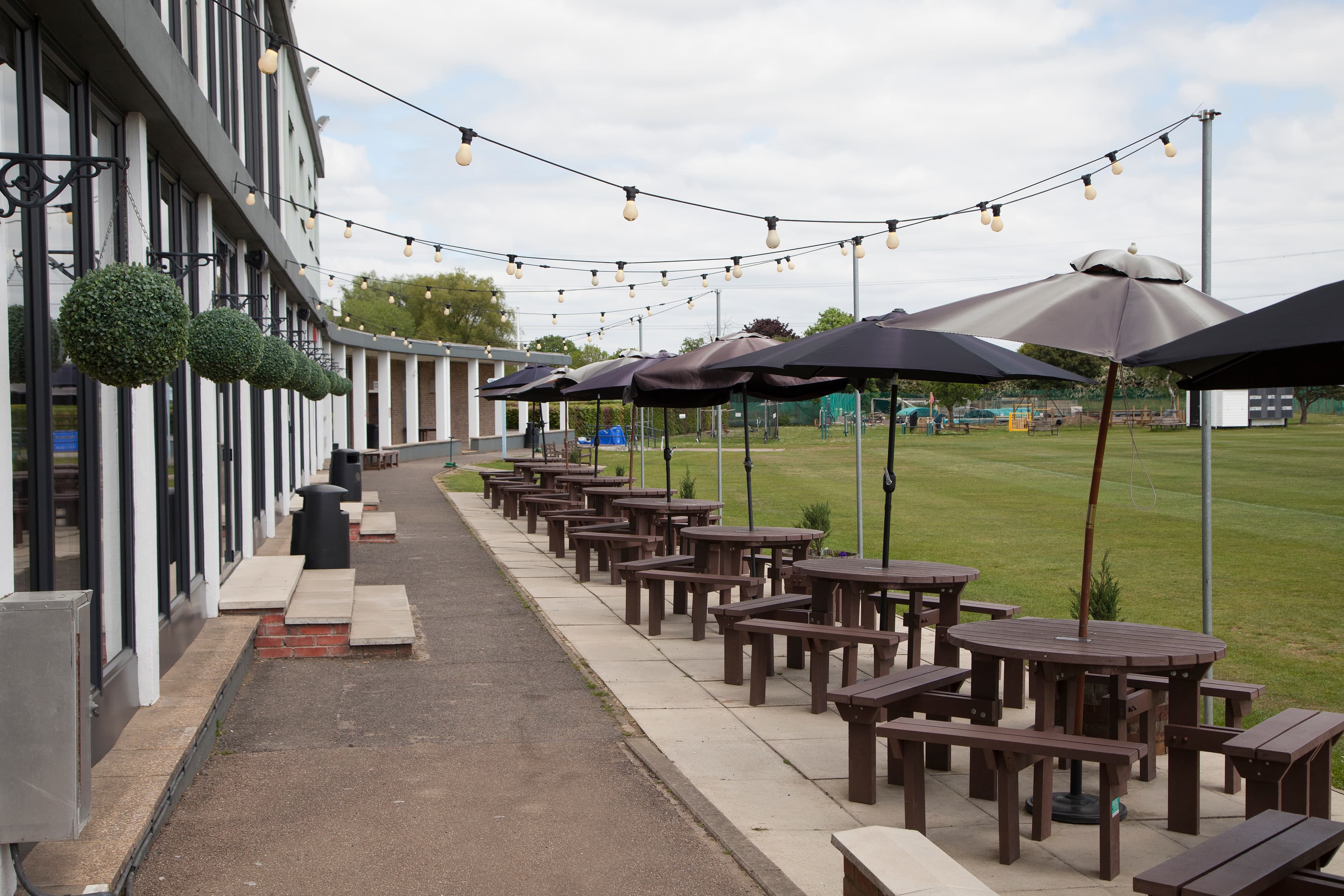 Outdoor terrace with dark wooden picnic benches, parasols and festoon lighting overlooking a sports field.