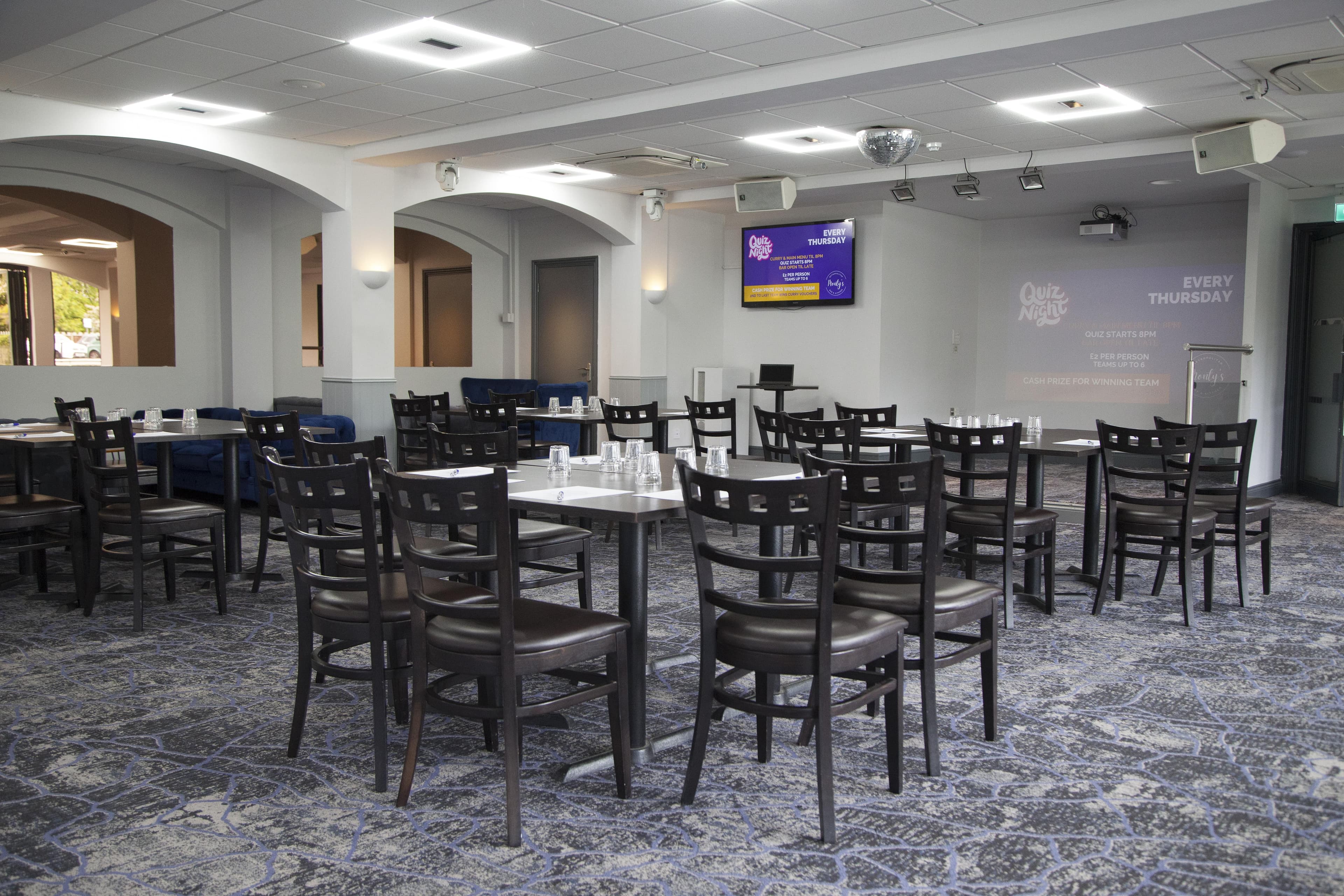 Carpeted function room with dark wood chairs, tables and quiz night screen projected on wall.