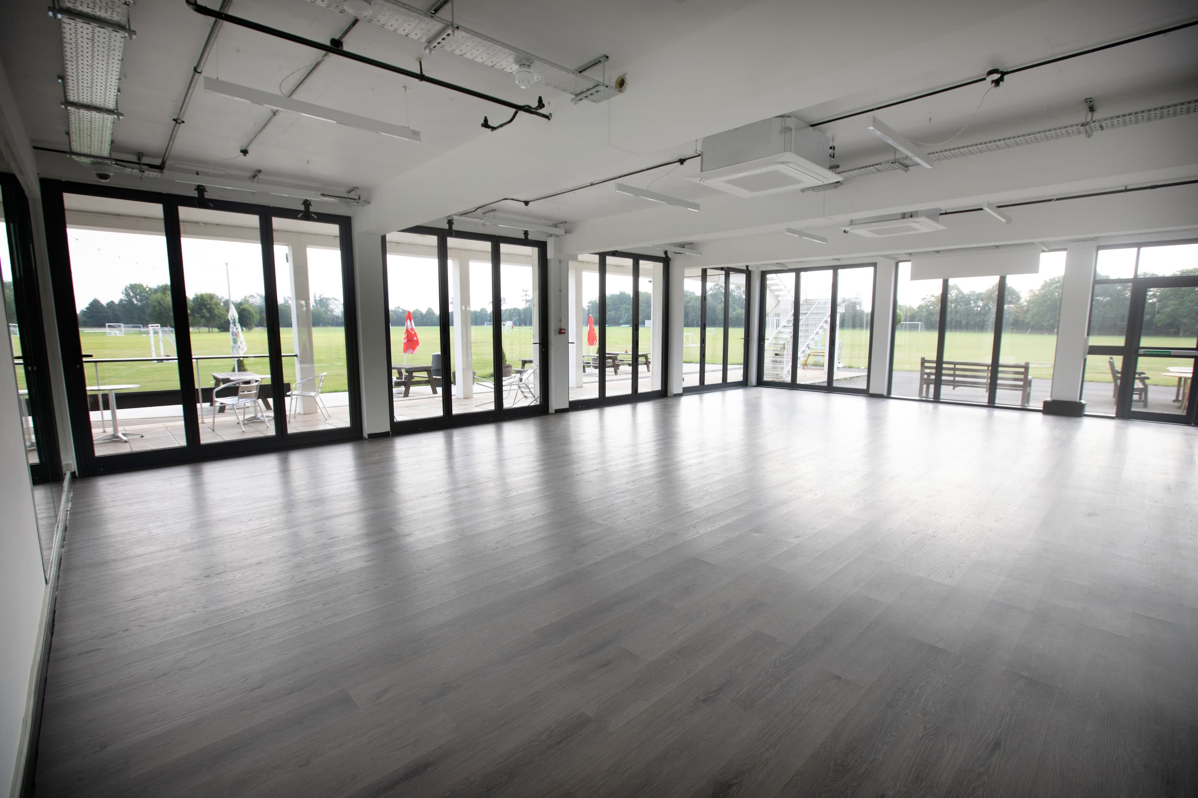 Bright empty function room with grey wood floor and floor-to-ceiling doors overlooking sports pitches.