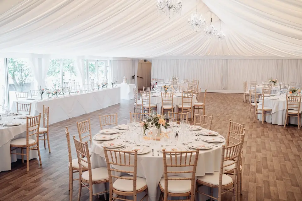 White draped marquee interior with round tables, chiavari chairs and floral centrepieces.