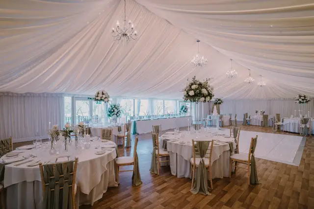 Draped marquee reception room with round tables, sage chair sashes and tall floral centrepieces.