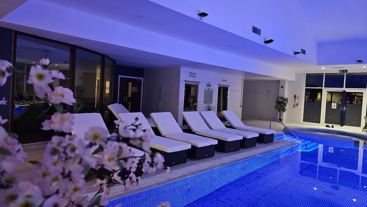 Indoor pool lit in blue with white sun loungers and steam room and sauna beyond.