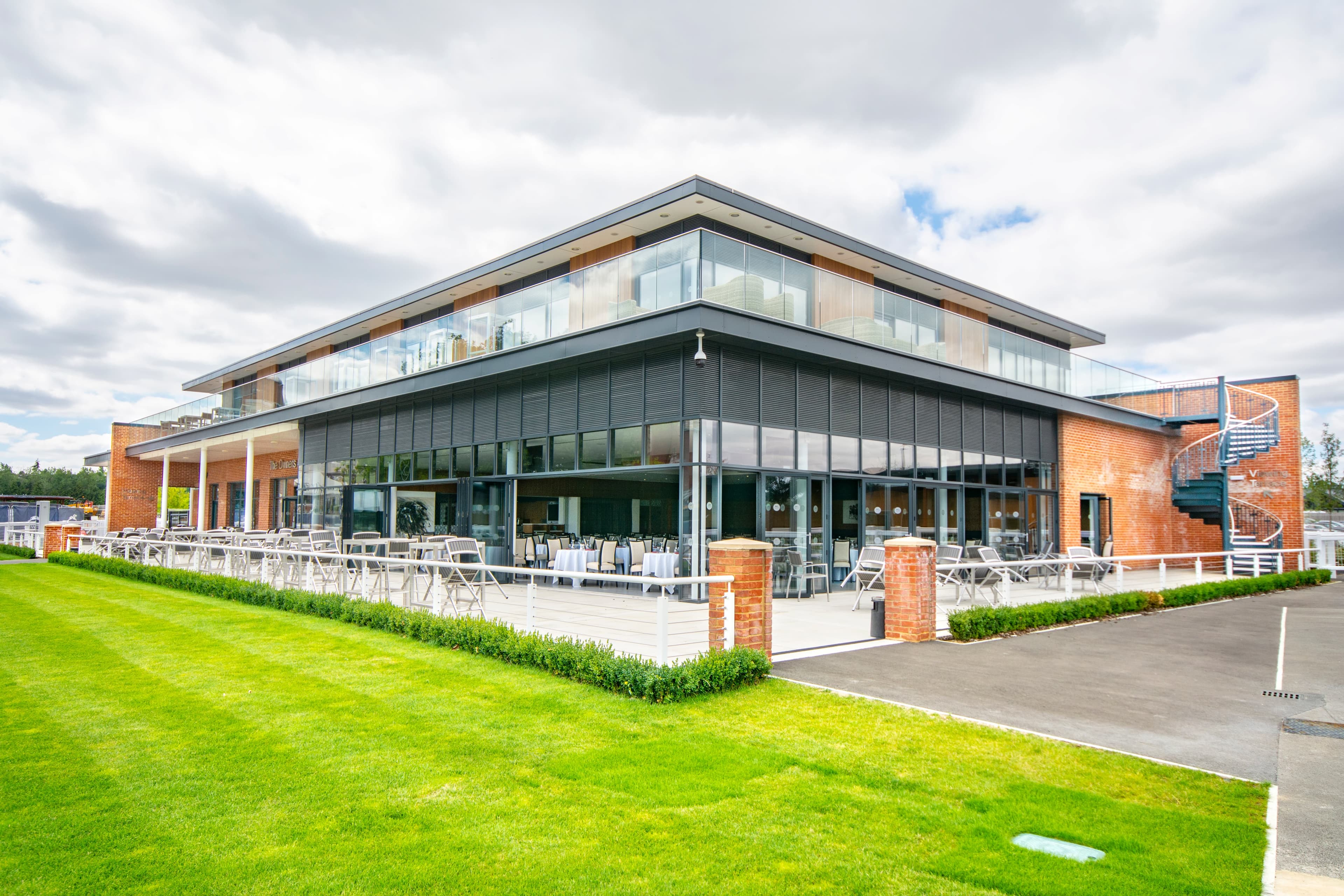 Modern two-storey glass and brick building with outdoor terrace seating and manicured lawn.