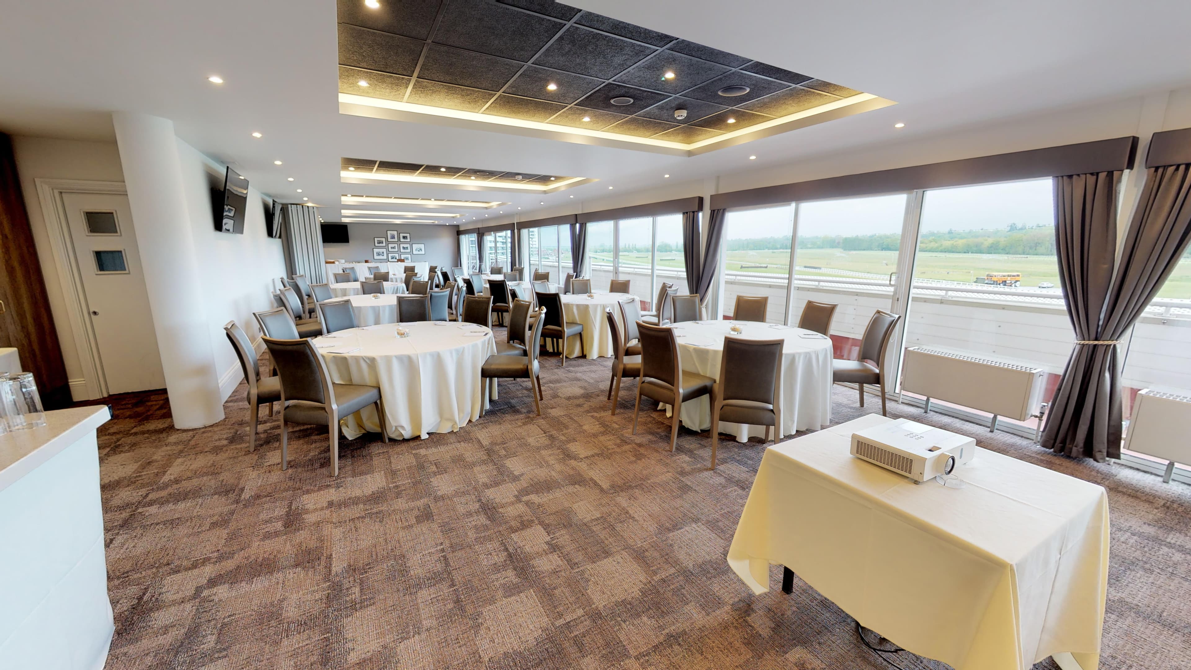 Bright function room with round tables, grey chairs and racecourse views beyond large windows.