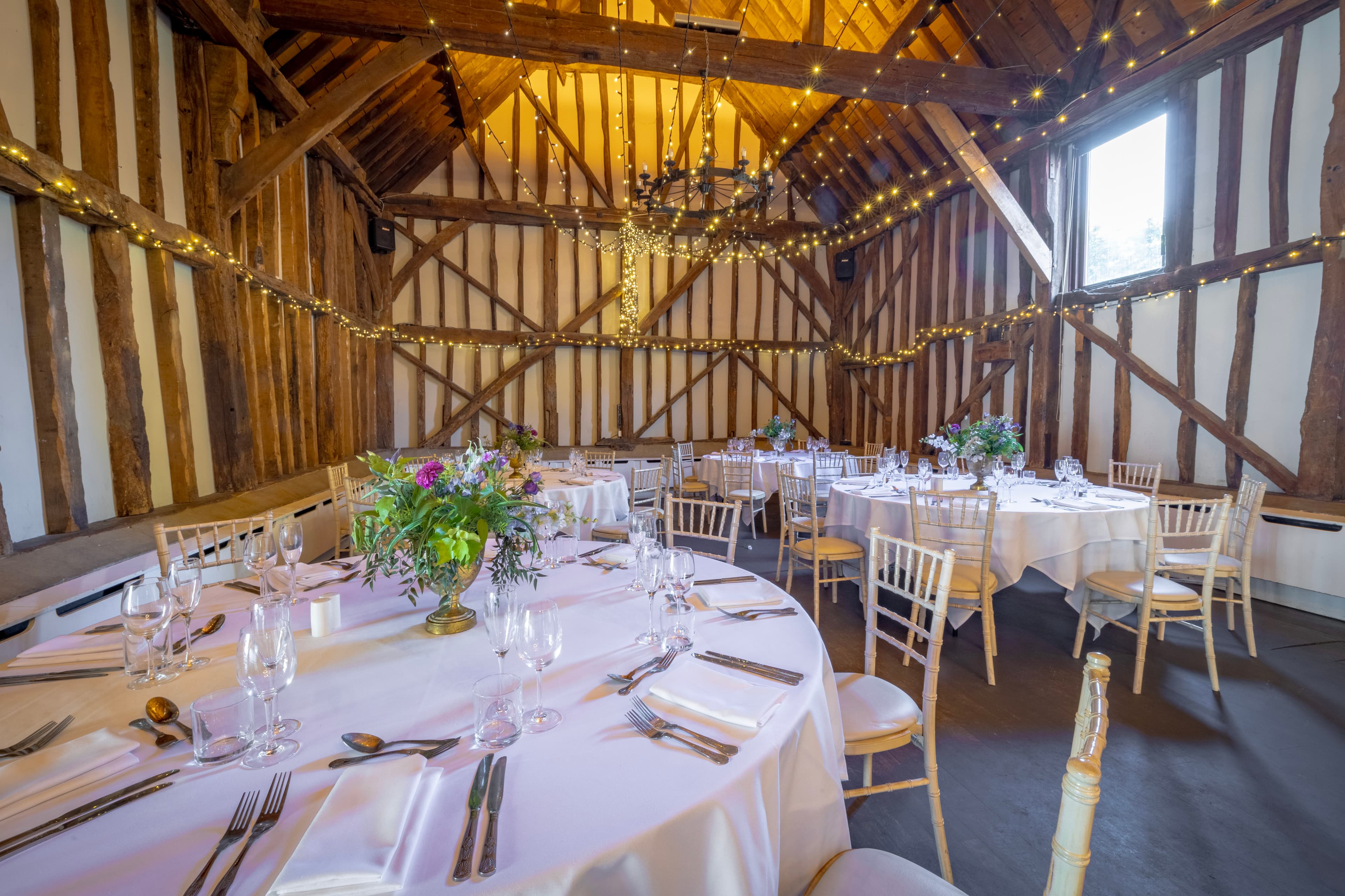 Timber-framed barn reception room with round tables, fairy lights and floral centrepieces.
