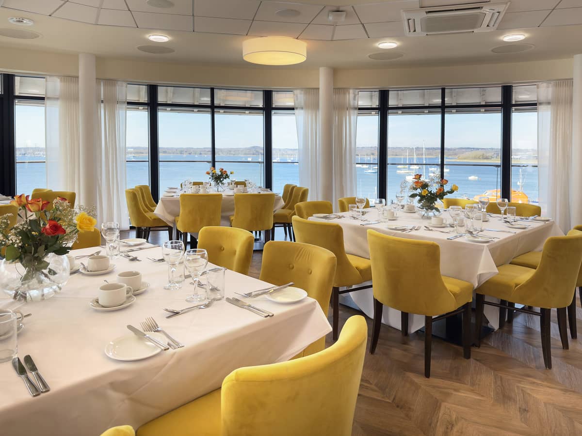Bright dining room with yellow chairs, laid tables and harbour views beyond floor-to-ceiling windows.
