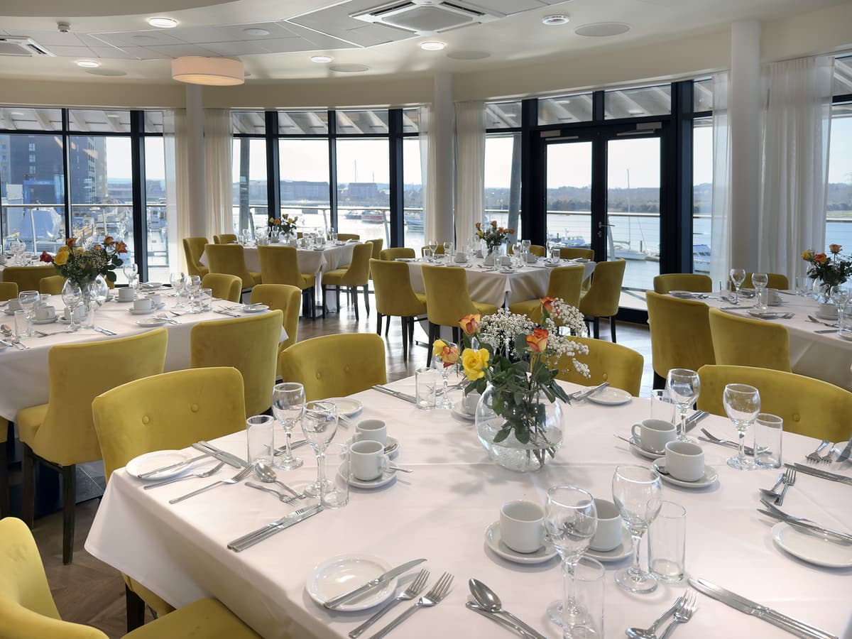 Bright waterside function room with round tables, yellow chairs and floral centrepieces.