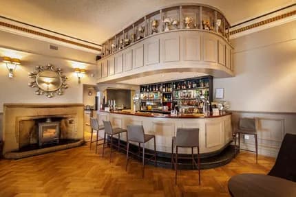 Curved hotel bar with high stools, backlit spirits shelves and a stone fireplace.